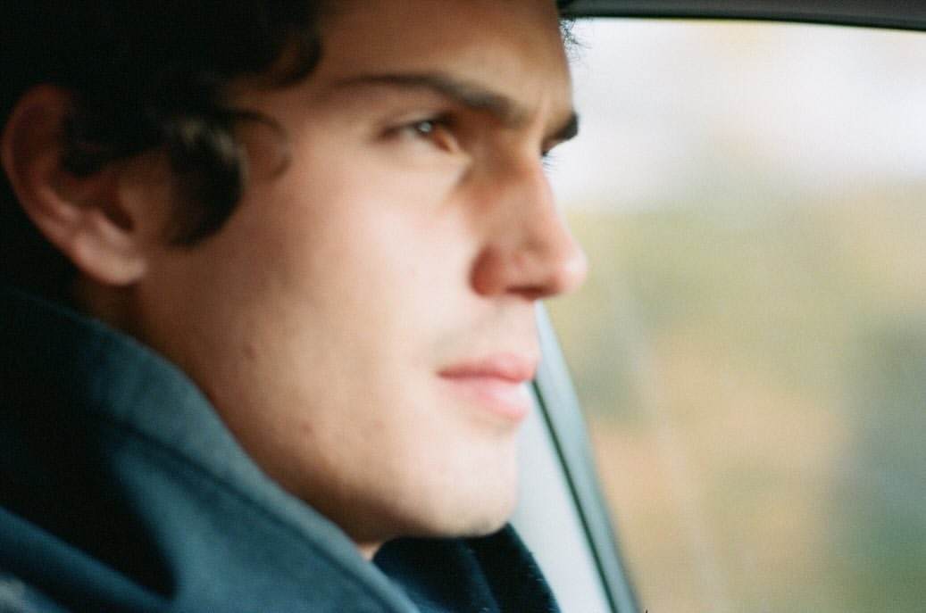 Close-up of a young adult man in profile, looking out of a car window with a serene expression and blurred background.
