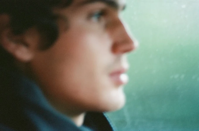 Close-up profile of a young man with dark wavy hair looking out a window with a green-toned background.