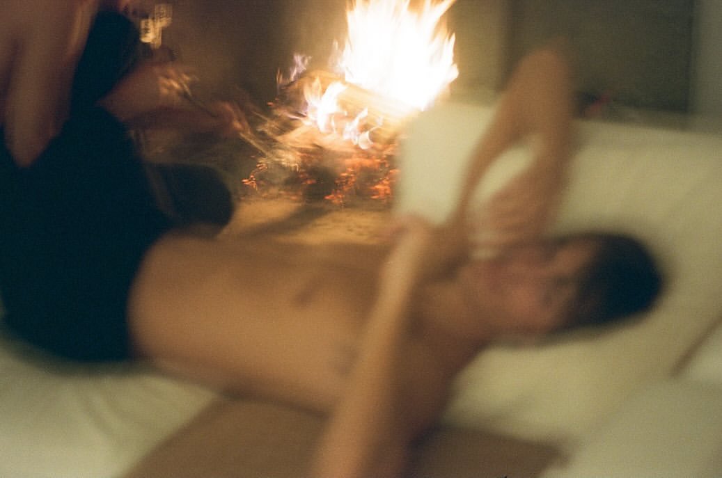 Blurred indoor scene with a shirtless man lying on a couch near a fireplace with a bright, flickering fire and warm lighting.