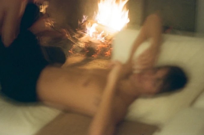Blurred indoor scene with a shirtless man lying on a couch near a fireplace with a bright, flickering fire and warm lighting.