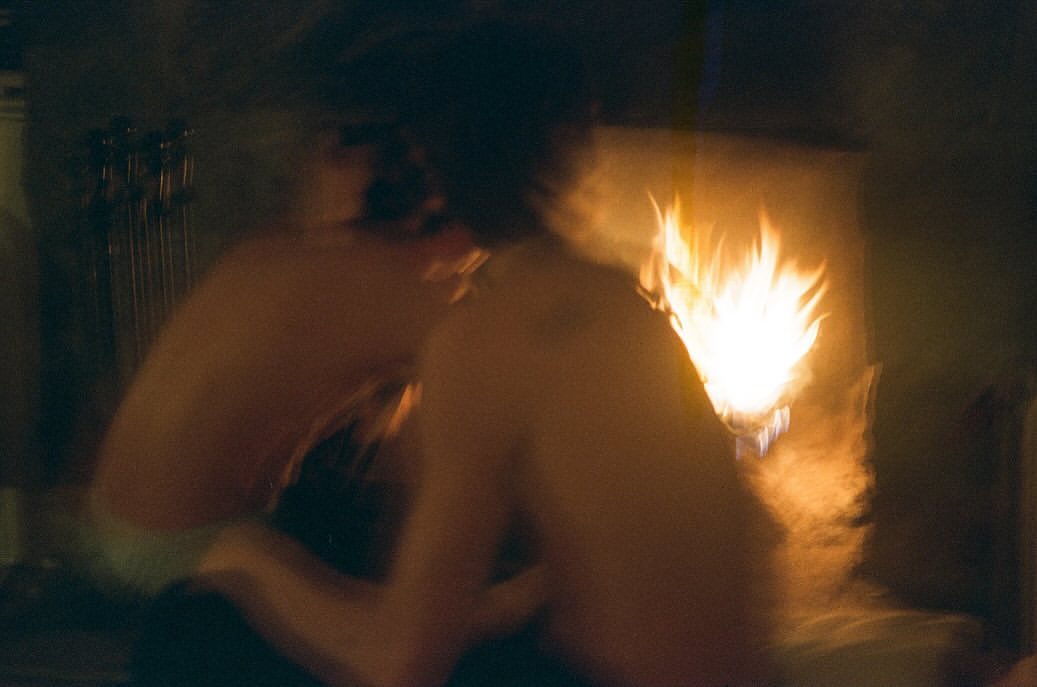 Two shirtless adults sit in a dark room lit by the warm glow of a fireplace, creating a cozy and intimate atmosphere.