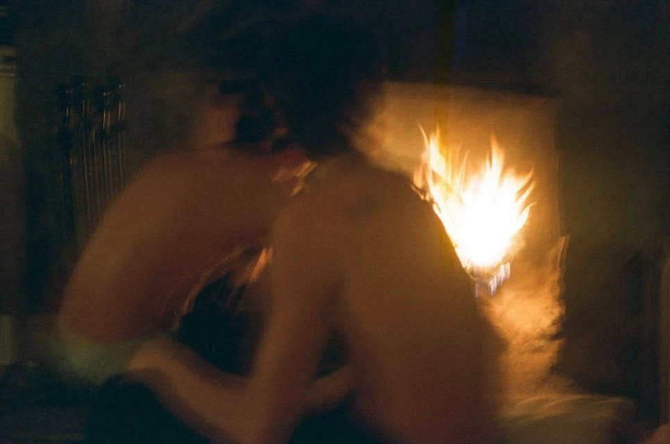Two shirtless adults sit in a dark room lit by the warm glow of a fireplace, creating a cozy and intimate atmosphere.