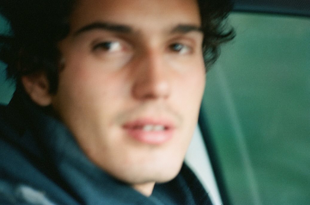 Close-up portrait of a young man with dark wavy hair and soft focus, seated inside a vehicle captured by Sebastian Faena.