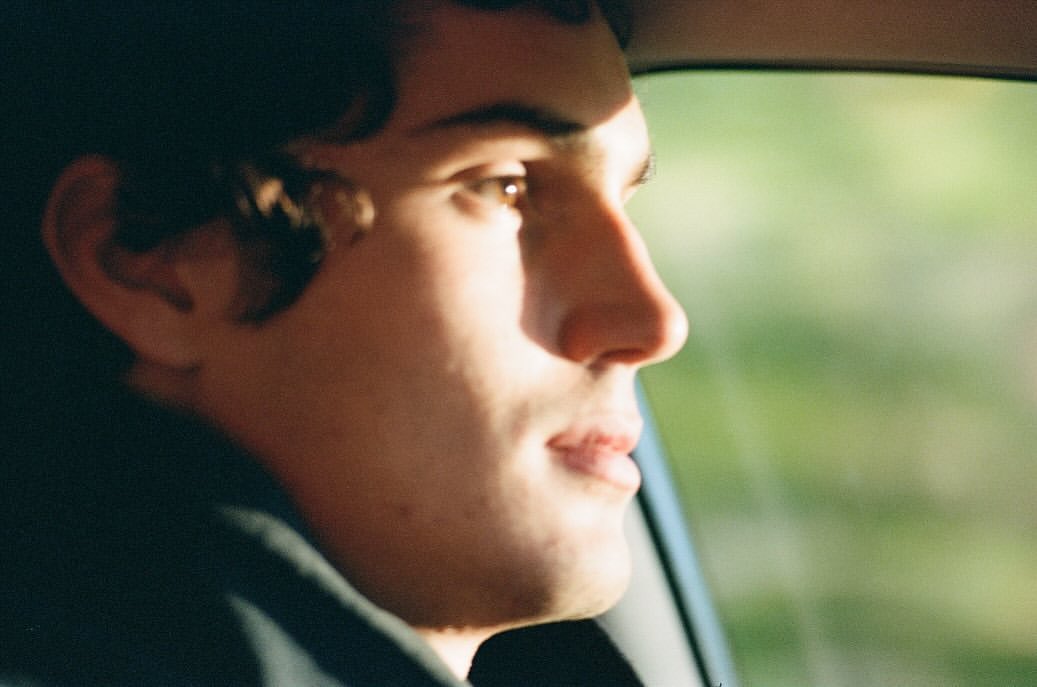 Close-up shot of a young adult man in profile inside a car. The background shows blurred green foliage through the car window.