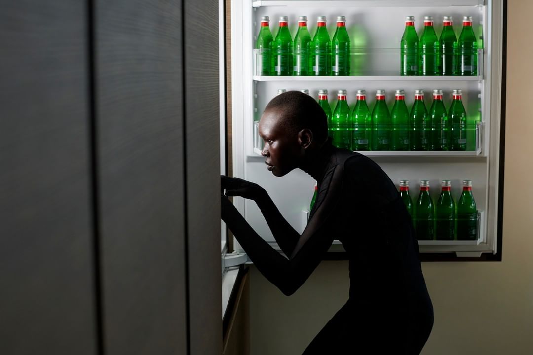Woman in black catsuit opens a fridge stocked with rows of green glass bottled drinks, creating a stark and modern, minimalist scene.
