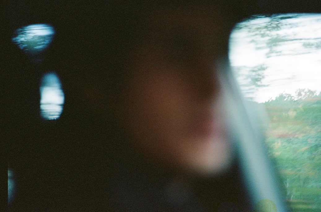 Blurred view of a person inside a vehicle, seen through the window, with blurry landscape visible in the background.