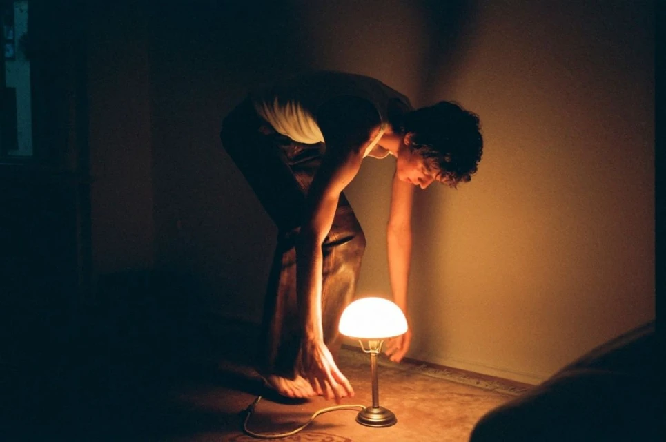 Indoor shot of a young adult looking down at a lit table lamp, casting warm light on the man and creating a cozy, intimate scene.