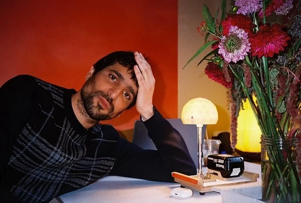 Indoor portrait of a man with flowers on a desk. He's resting his head on his hand, wearing a sweater, with flowers and desk lamp nearby.