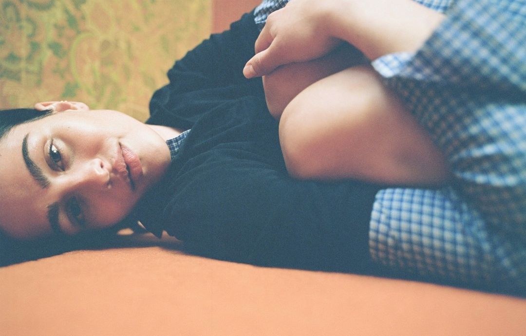 Portrait of a woman lying down wearing a blue and white checkered shirt, looking at the camera, on an orange surface.