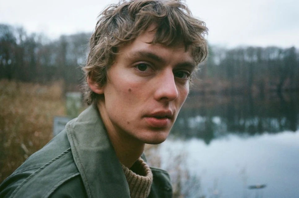 Close-up shot of a fair-skinned young adult with curly brown hair and an olive jacket against a blurred lake and forest backdrop.