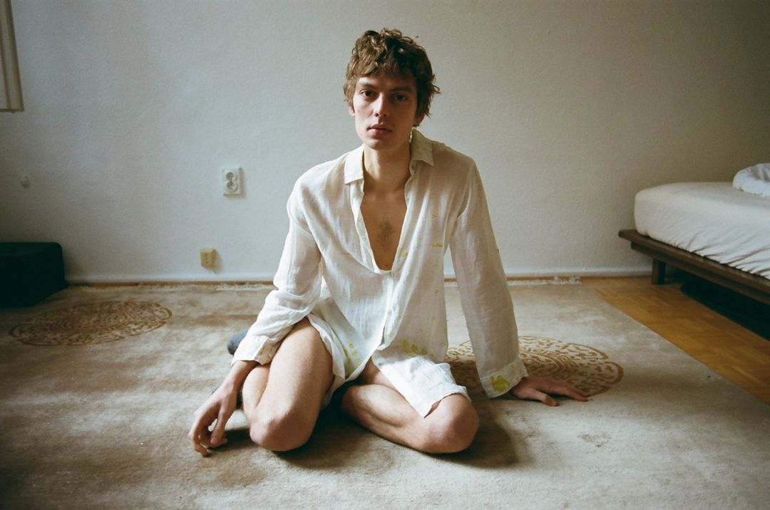 Young man sits cross-legged on a rug in a white shirt. Soft, diffused light fills the room. Bed and wall in the background add context.