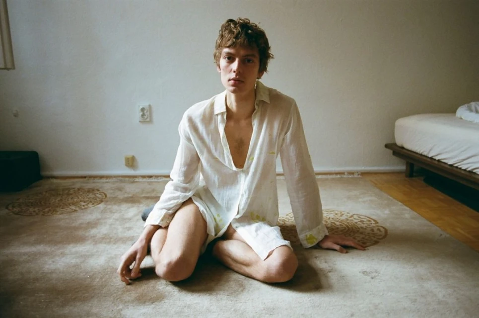Young man sits cross-legged on a rug in a white shirt. Soft, diffused light fills the room. Bed and wall in the background add context.