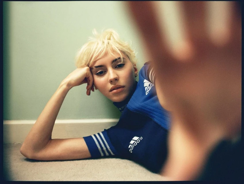Blonde woman in Adidas sports top poses indoors with hand close to camera. The background is a muted green wall and neutral carpet.