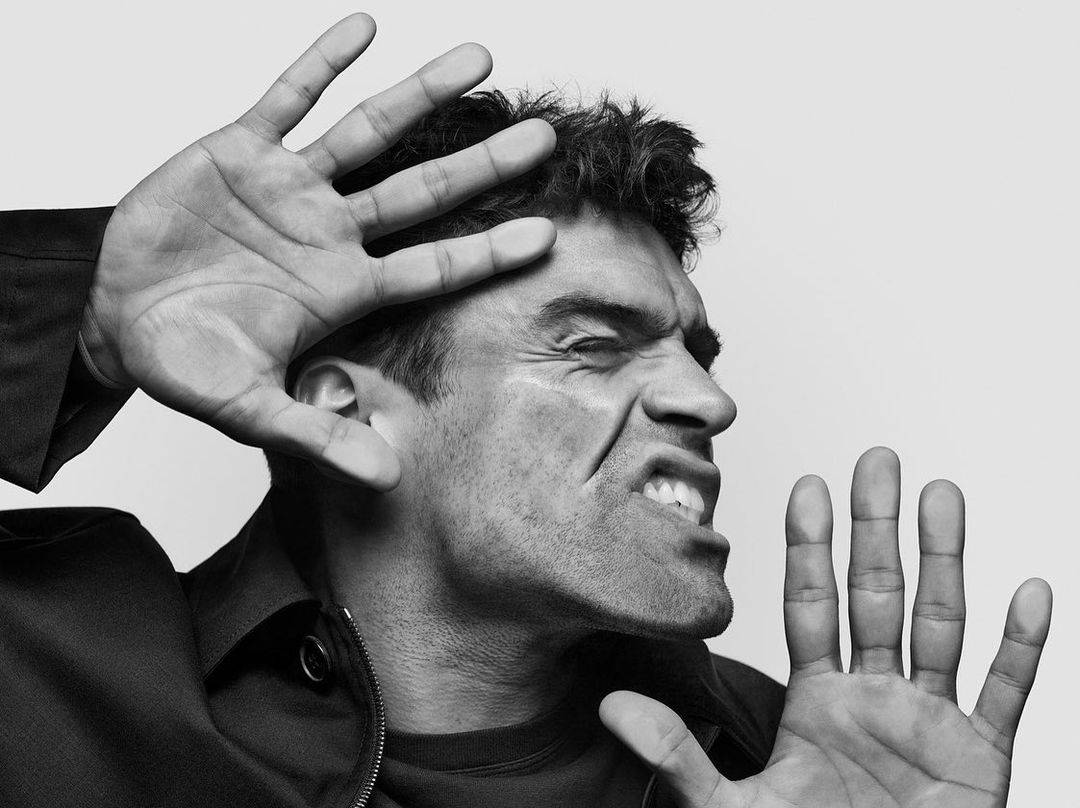 Black and white portrait of adult male with hands up and a distressed expression on a plain background with a dark jacket.