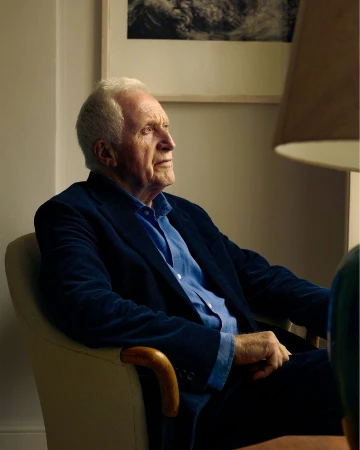 Elderly man with white hair seated in an armchair wearing a blue velvet blazer and blue button-down shirt by Sebastian Nevols