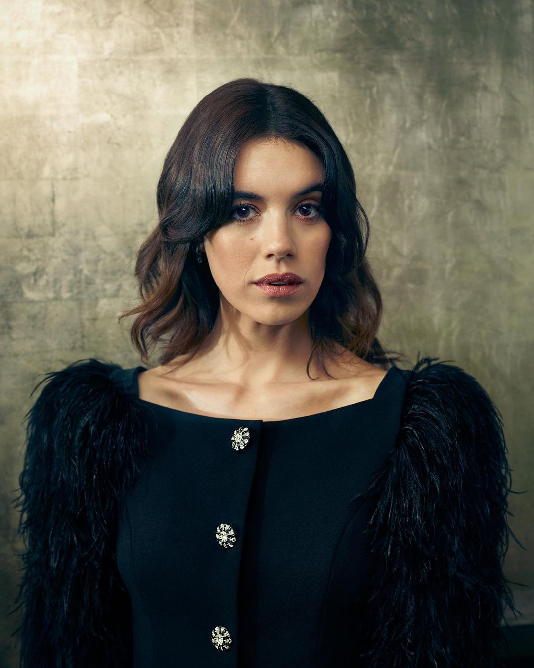 Portrait of a young woman with dark hair wearing a black dress with feather sleeves and decorative buttons against a textured gold background.