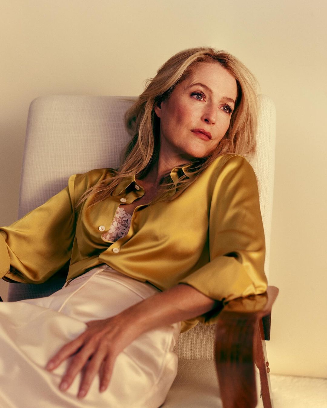 Blonde woman in a golden blouse sitting in a chair in a studio. Natural light illuminating her features and blonde hair.
