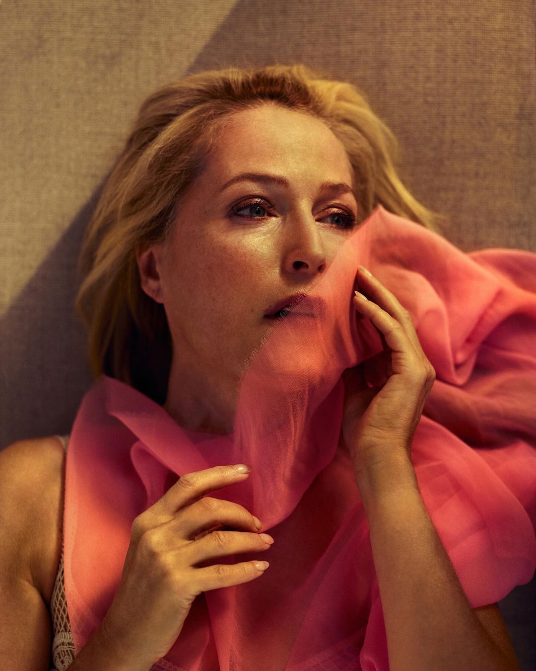 Elegant portrait of adult female lying down, holding pink fabric over her face in warm light, conveying a serene and artistic mood.