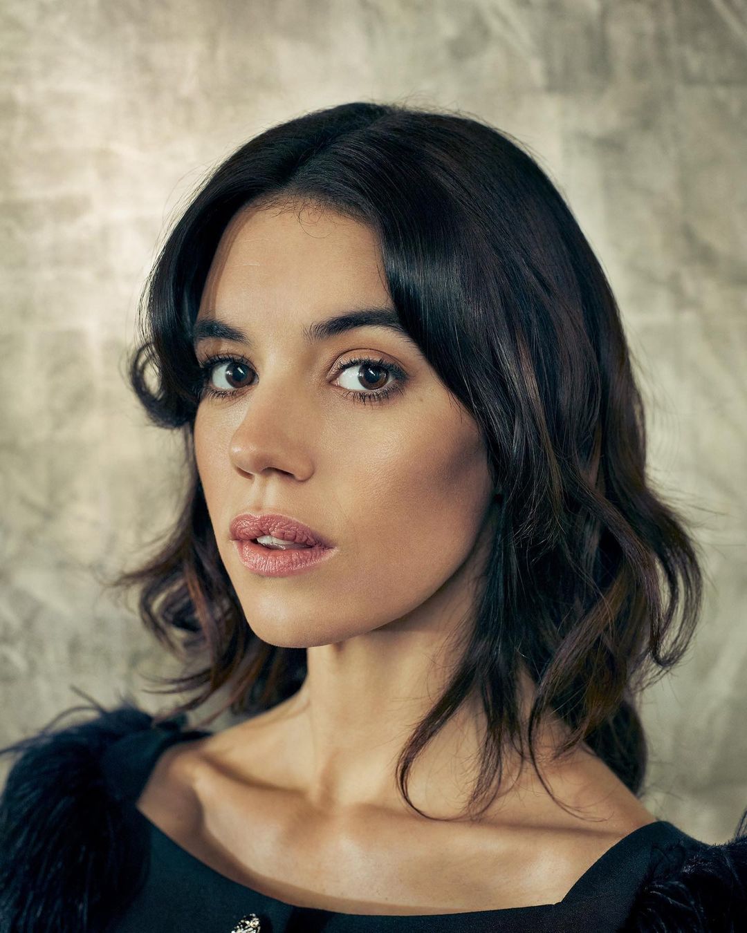 Portrait of an adult woman with dark wavy hair looking at the camera against a neutral background in a close-up shot.
