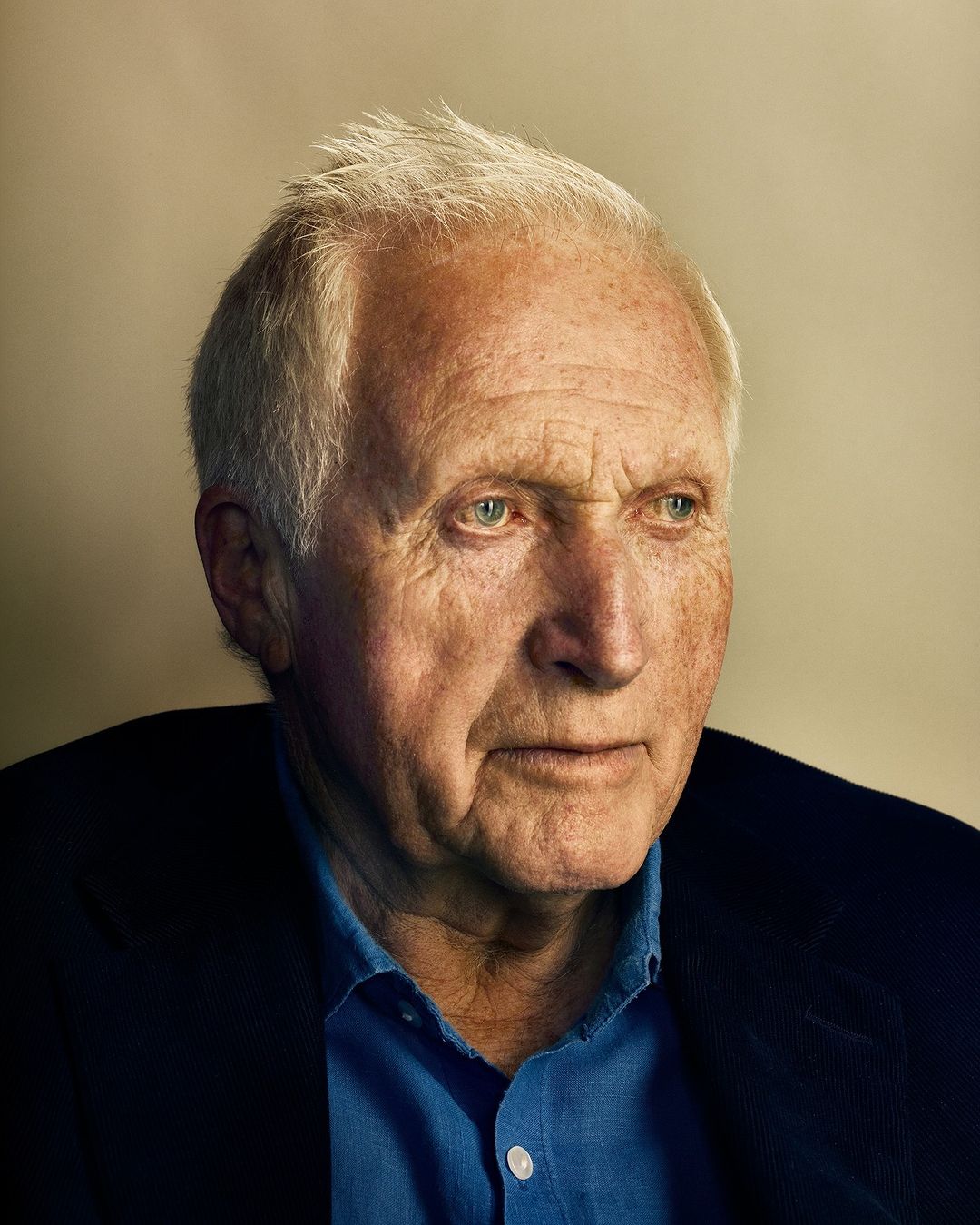 Close-up portrait of an elderly Caucasian man with gray hair wearing a blue shirt and dark jacket, looking off to the side.