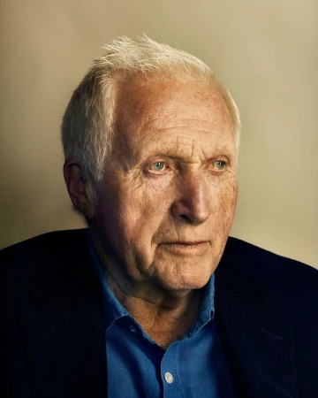Close-up portrait of an elderly Caucasian man with gray hair wearing a blue shirt and dark jacket, looking off to the side.