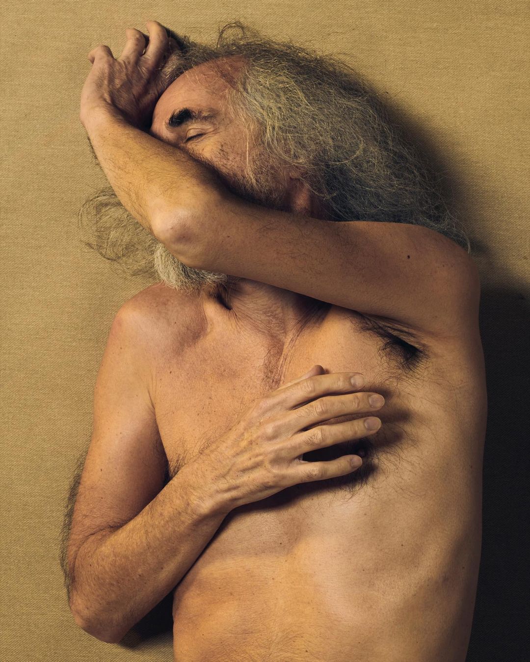 Topless adult male with long gray hair covers his face with his hands. He lies against a plain tan background in a close-up studio shot.