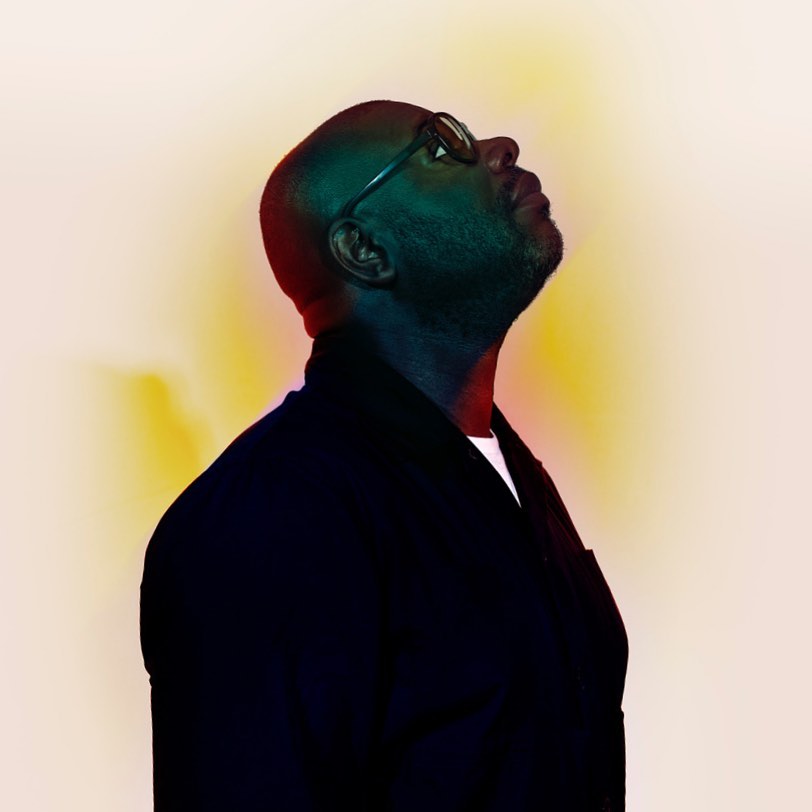Portrait of an adult male with shaved head, glasses, and dark jacket looking up against a diffused gradient background.