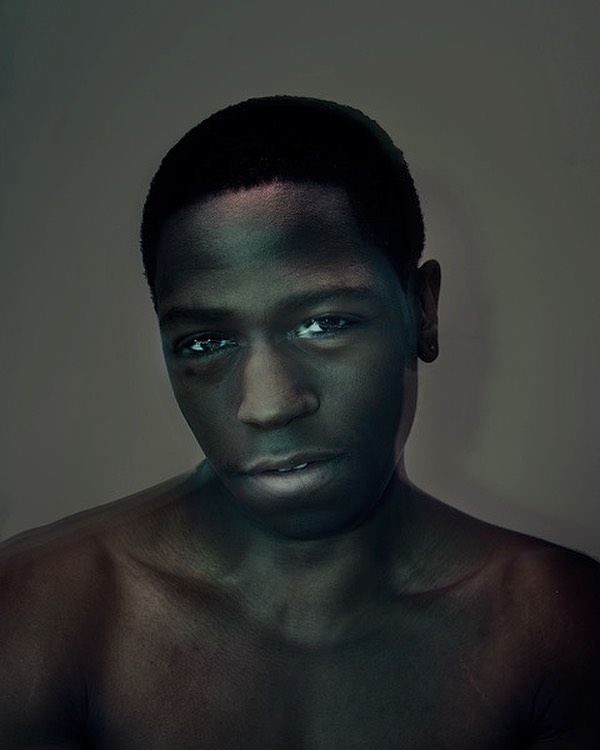 Portrait of a young African American male model with short hair, gazing into the camera with a neutral facial expression in a studio setting.