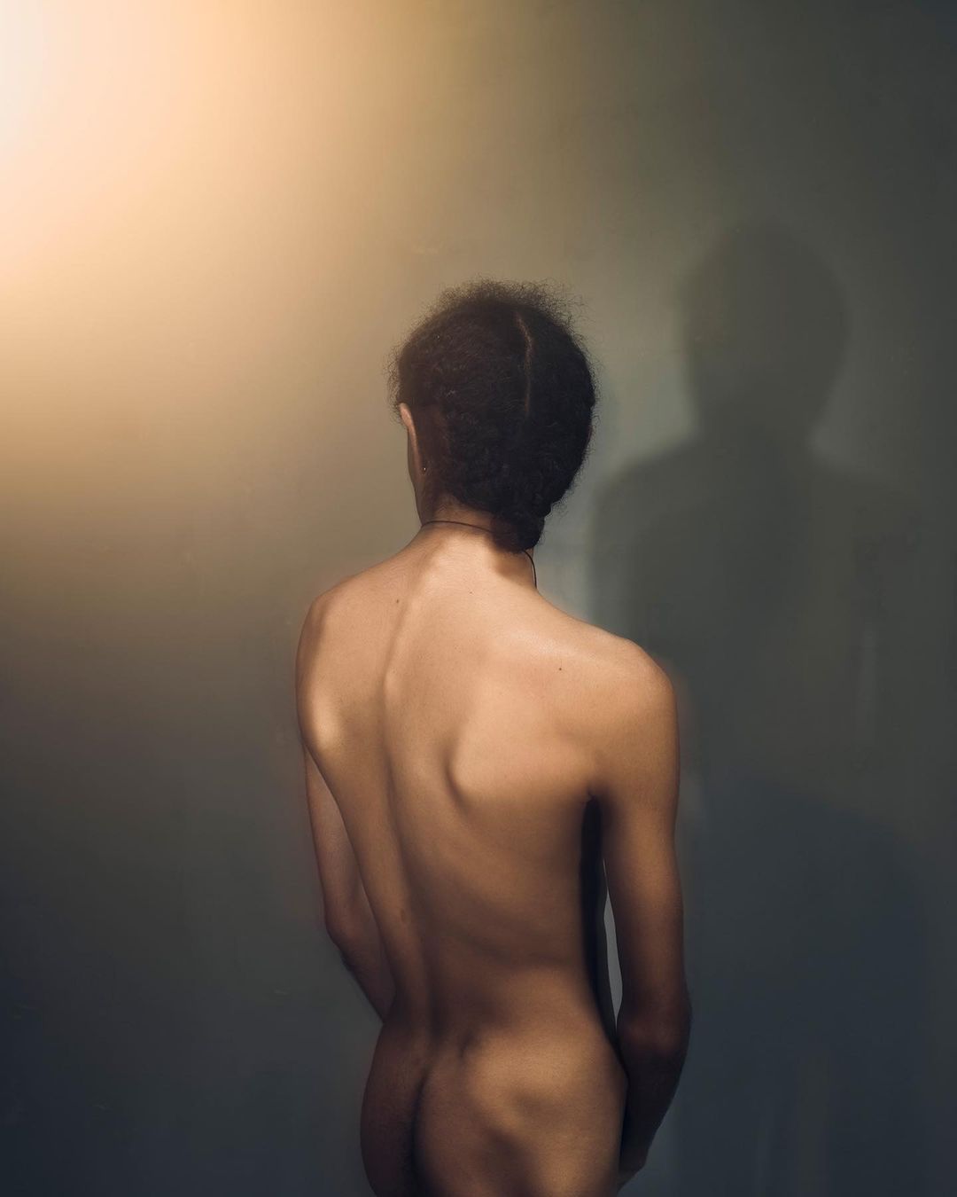 Back view of a shirtless adult with dark curly hair, gazing at a wall with a shadow, evoking a sense of introspection and solitude.