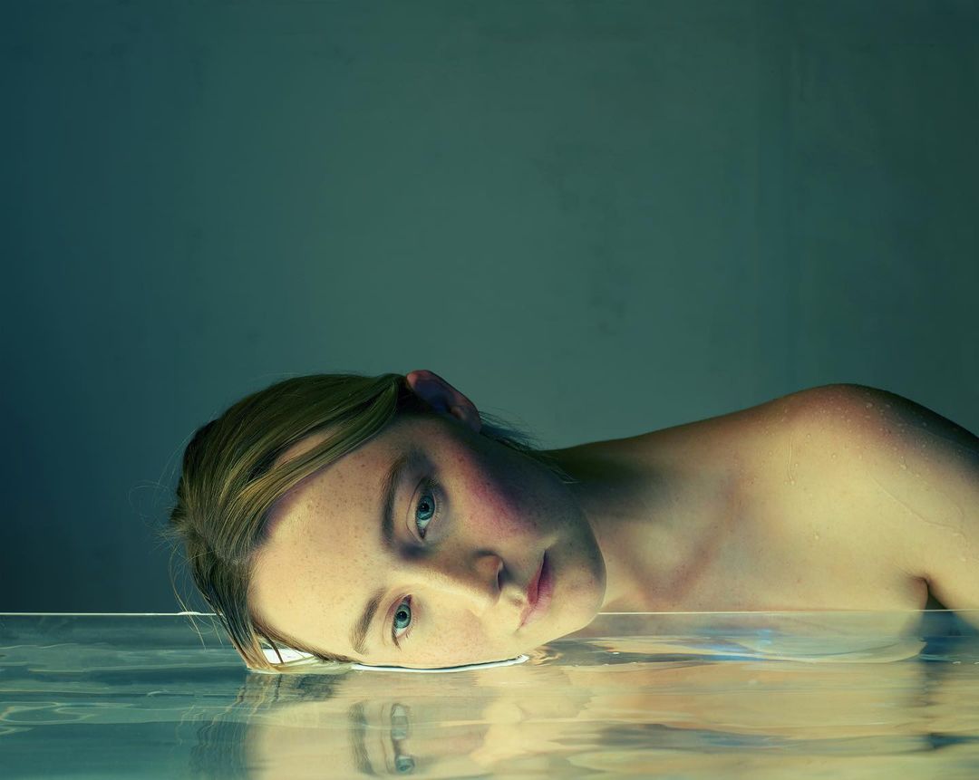 Young woman with blonde hair rests her head on a clear, reflective surface with a soft blue background and diffused lighting.
