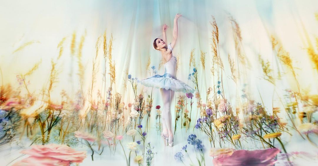 Ballerina in white tutu poses en pointe among wild flowers and tall grasses under soft, diffused light creating an ethereal effect.