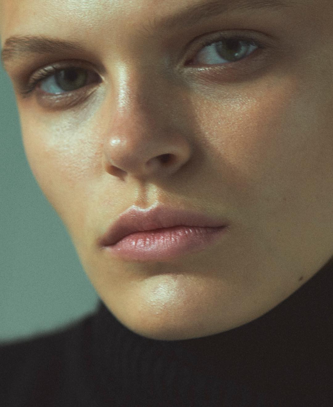 Close-up portrait of an adult female with fair skin, light eyes, and glossy lips wearing a black turtleneck in a soft light setting.
