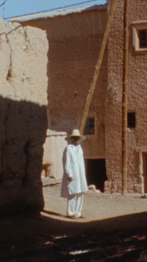 Person in white suit and hat standing on dirt road in front of stone buildings in an outdoor, sunlit, daytime environment.