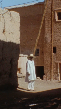 Person in white suit and hat standing on dirt road in front of stone buildings in an outdoor, sunlit, daytime environment.