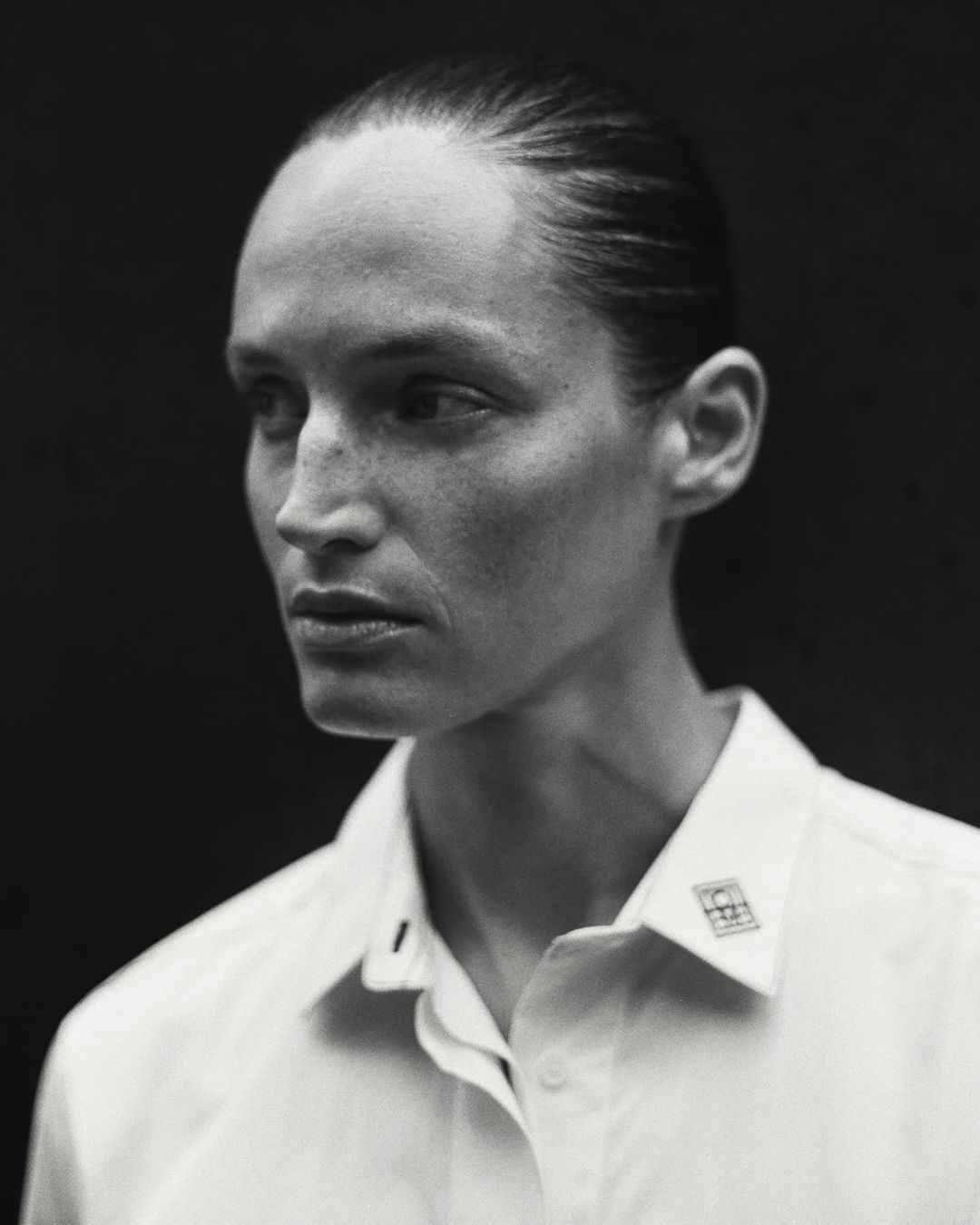 Monochrome portrait of an adult woman with slicked-back hair wearing a white button-down shirt against a dark background in a photo studio.
