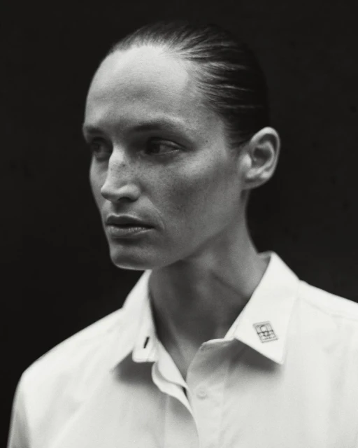 Monochrome portrait of an adult woman with slicked-back hair wearing a white button-down shirt against a dark background in a photo studio.