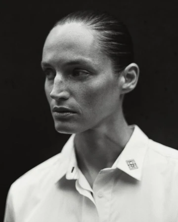 Monochrome portrait of an adult woman with slicked-back hair wearing a white button-down shirt against a dark background in a photo studio.