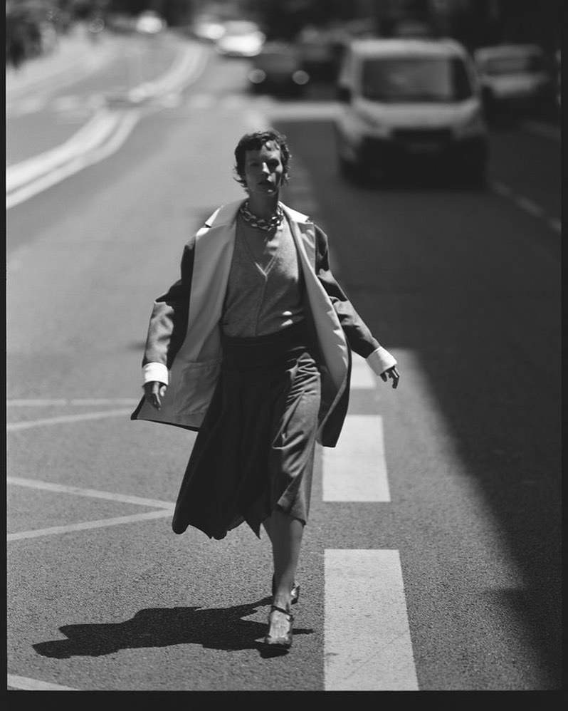 Model walking across a city street wearing a coat draped over shoulders, captured in black and white photography.