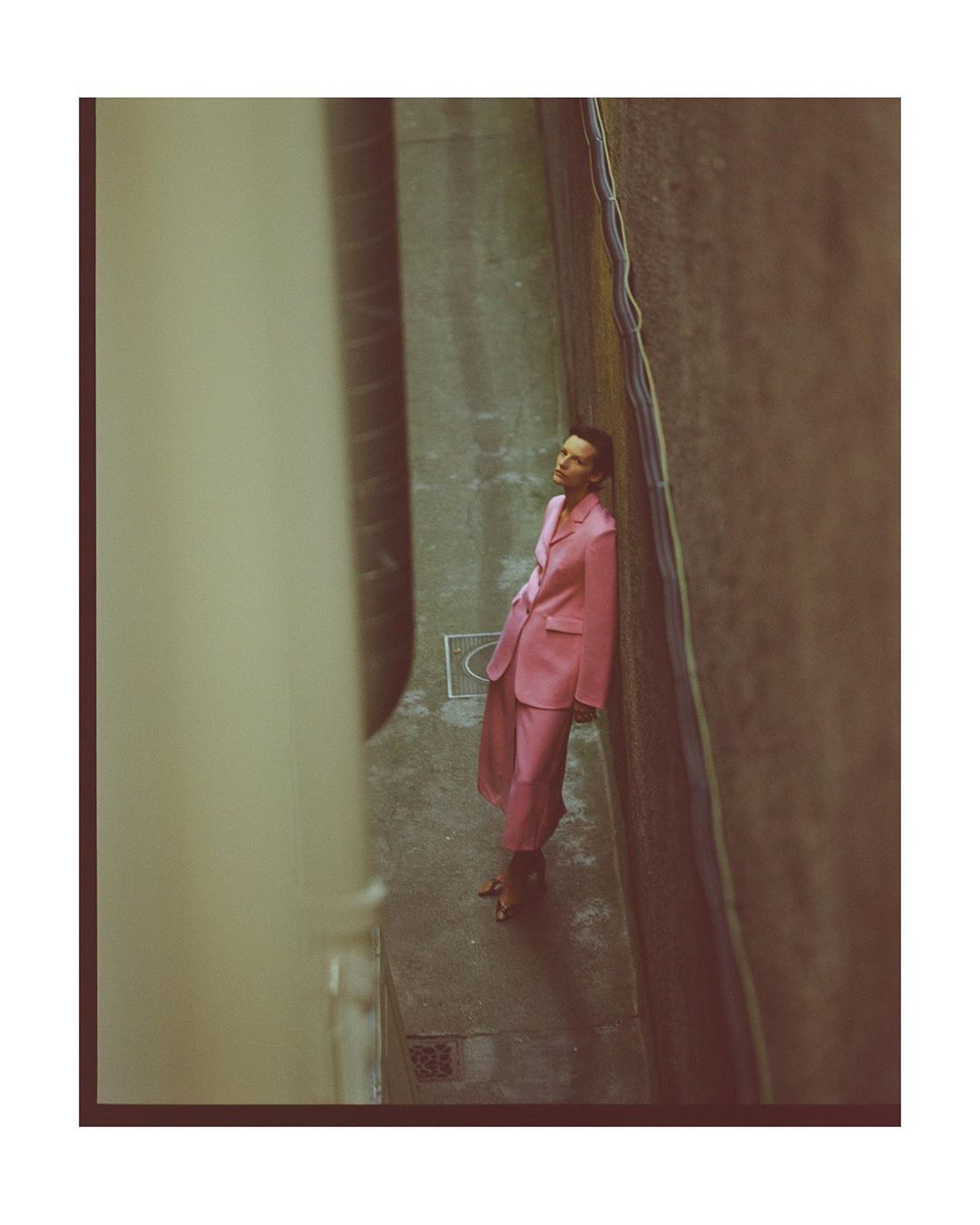 Woman in a pink suit leans against a concrete wall in a narrow alleyway with overhead pipes, creating an editorial fashion shot.