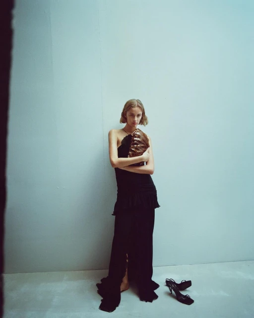 Fashion portrait of a woman in a black dress holding a brown purse, posed in front of a blue wall with black high heels on the floor.