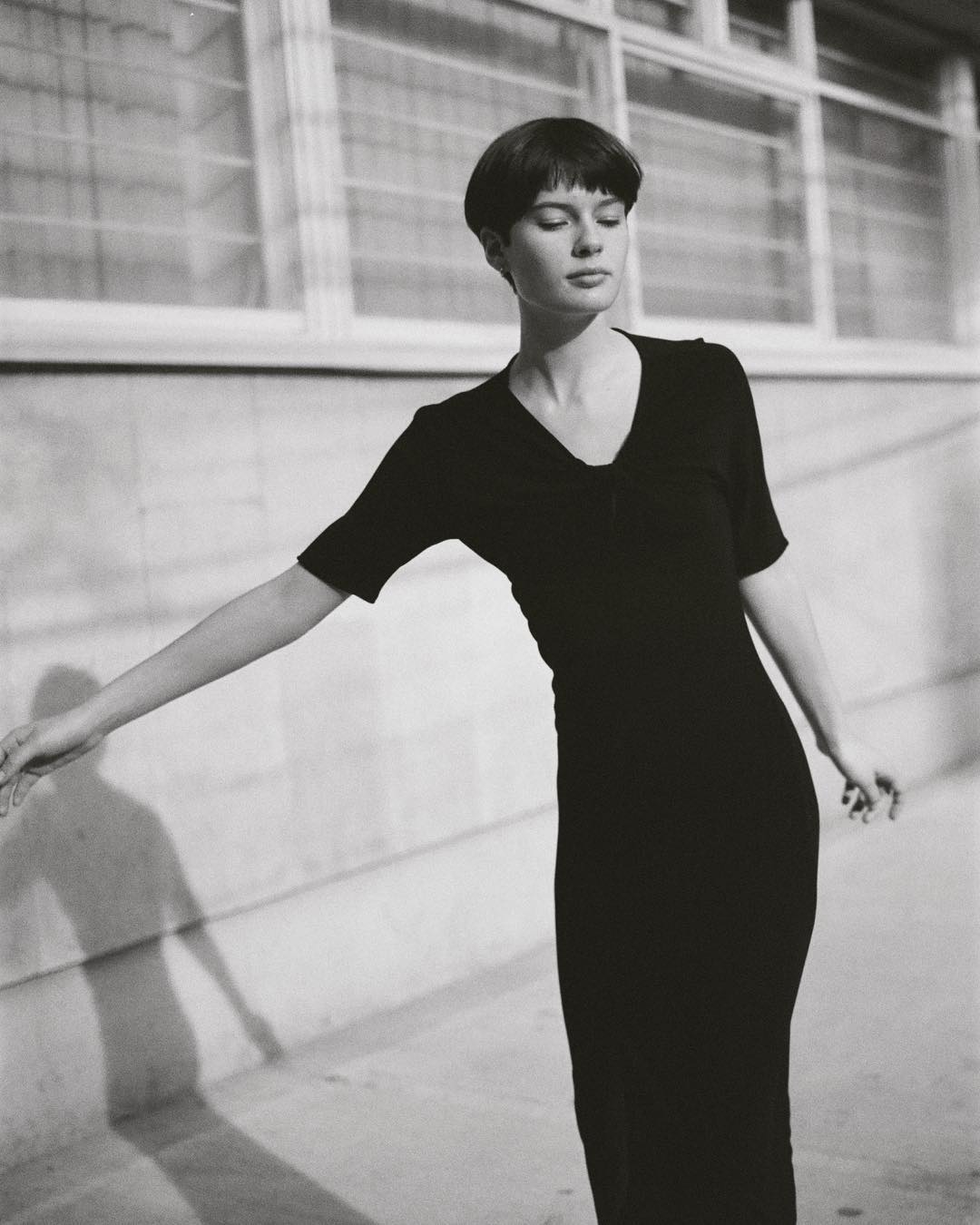 Elegant black and white portrait of a woman with a bowl cut wearing a black dress, posing outdoors with a concrete building in the background.