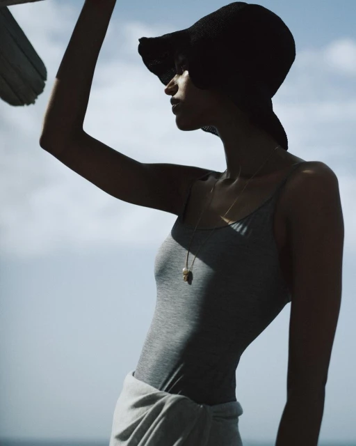 Woman in gray tank top and black hat poses against a sky with clouds. Necklace visible. Soft, natural lighting.