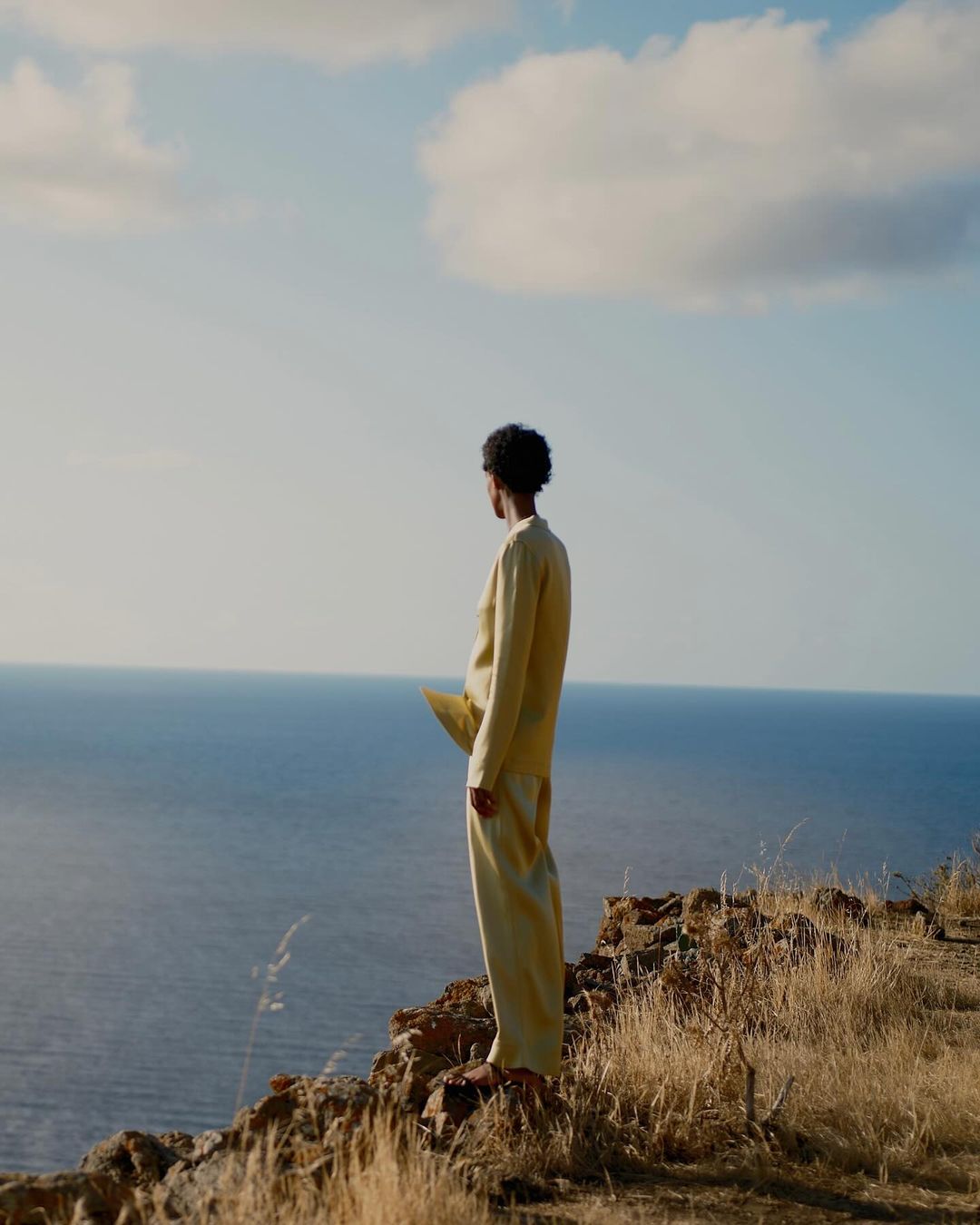 Person in yellow clothing stands on a cliff overlooking the ocean under a sunny sky, enjoying the scenic landscape. Landscape orientation.