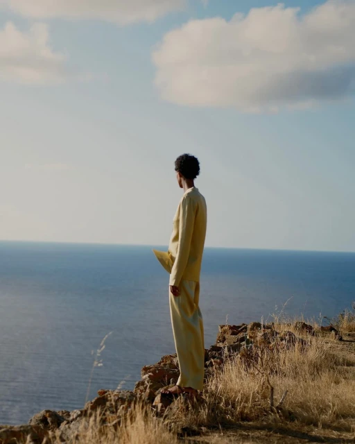 Person in yellow clothing stands on a cliff overlooking the ocean under a sunny sky, enjoying the scenic landscape. Landscape orientation.
