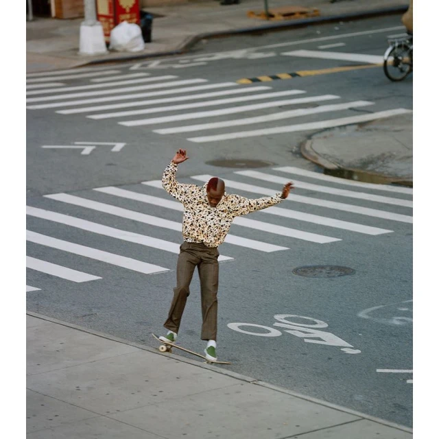 Young adult rides skateboard along crosswalk in urban setting, wearing printed top and brown pants, captured in a high-angle shot.