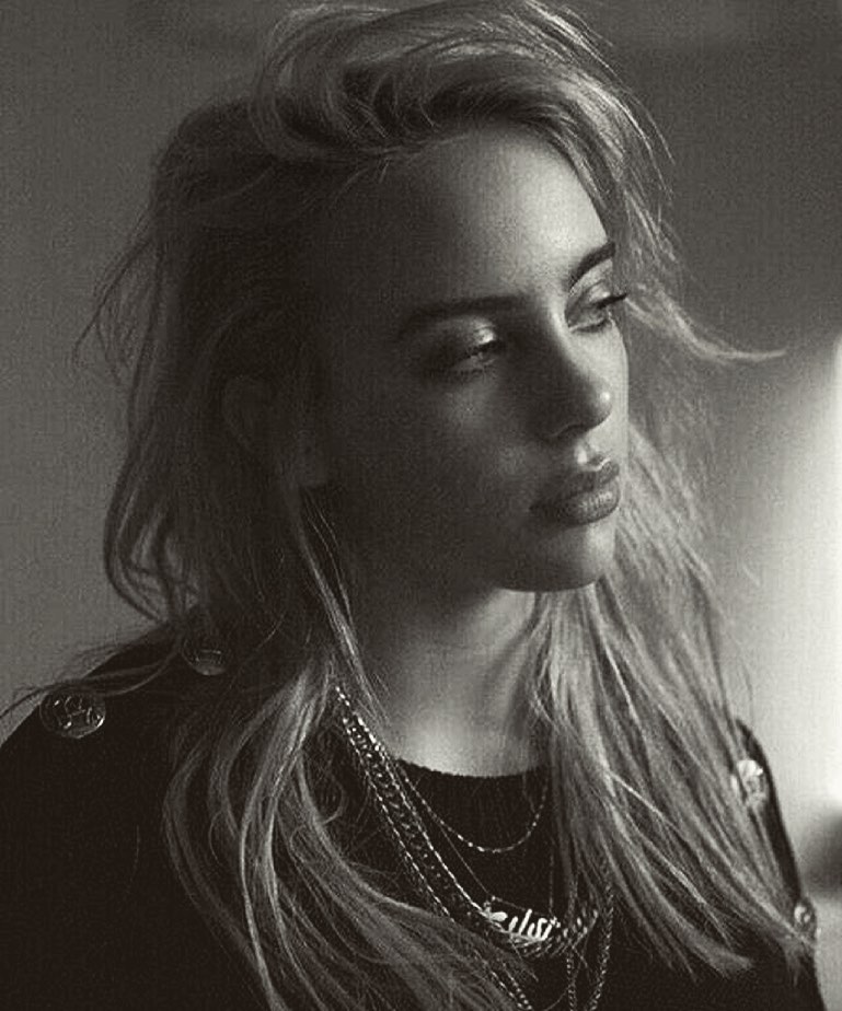 Portrait of Billie Eilish with layered chains wearing a dark top against a plain background photographed by Sebastian Sabal-bruce.