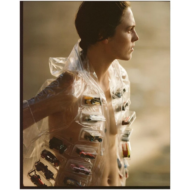 Profile view of a person wearing a clear plastic jacket filled with toy cars, set against a hazy brown background in a fashion shot.