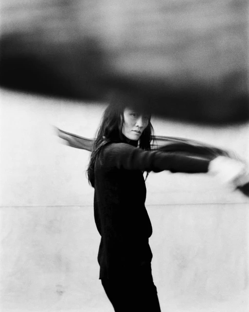 Black and white portrait of an adult woman with long dark hair, wearing a dark turtleneck shirt, in blurred motion against a light background.