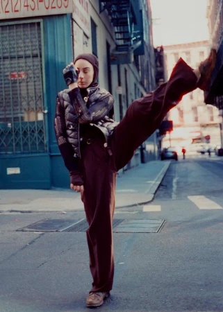 Woman in puffy jacket and brown pants balances with a high kick on a city street with vintage-style buildings and muted lighting.