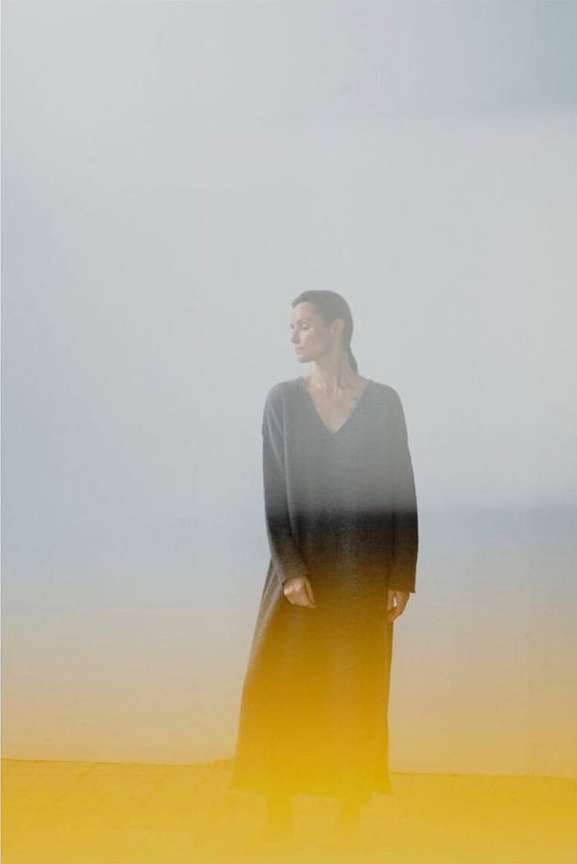 A woman in a long-sleeve maxi dress poses against a foggy grey background with yellow tint, looking off to the side in a fashion editorial.